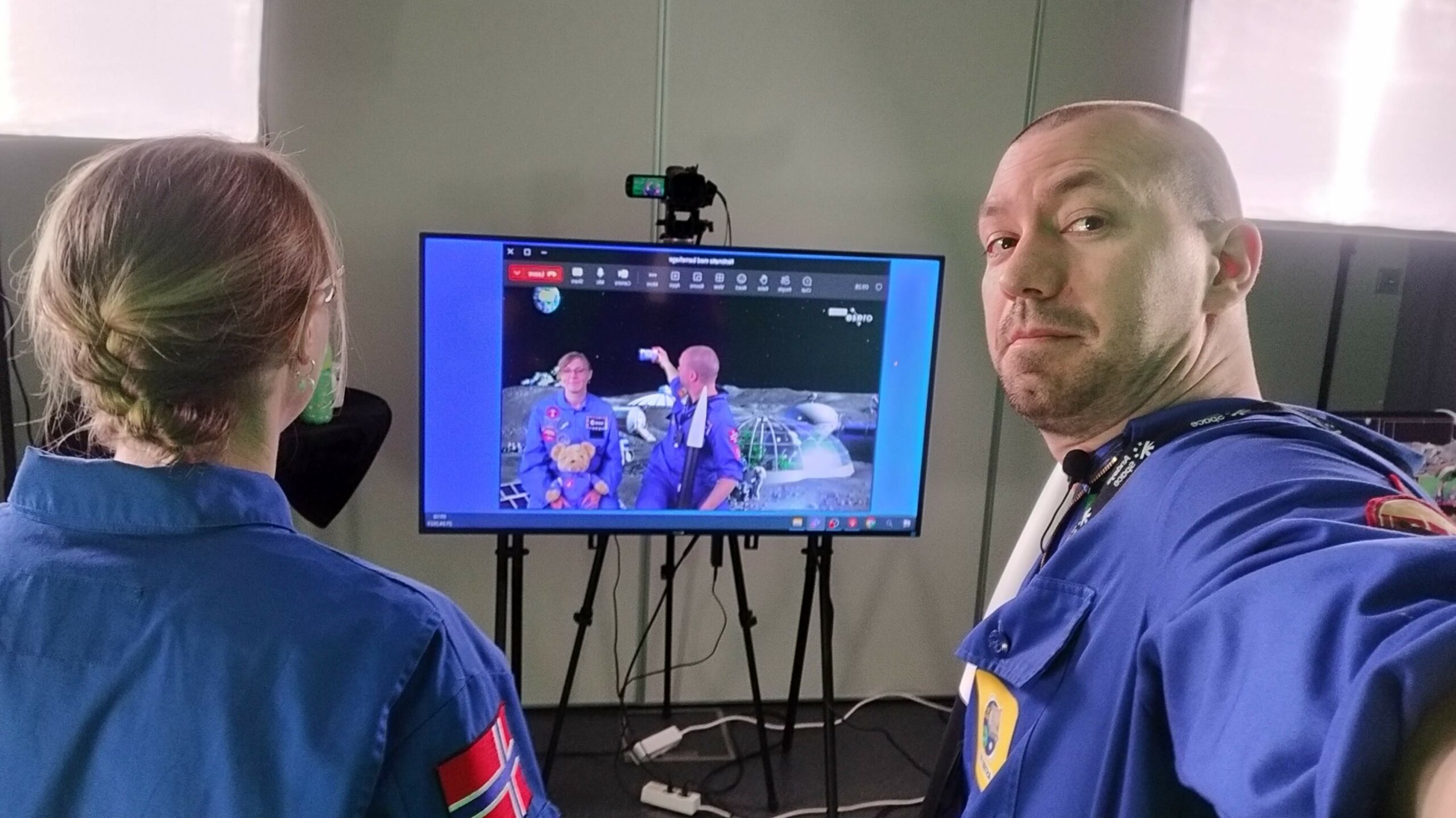 To personer i blå flight suit foran en tv og kamera i et studio.