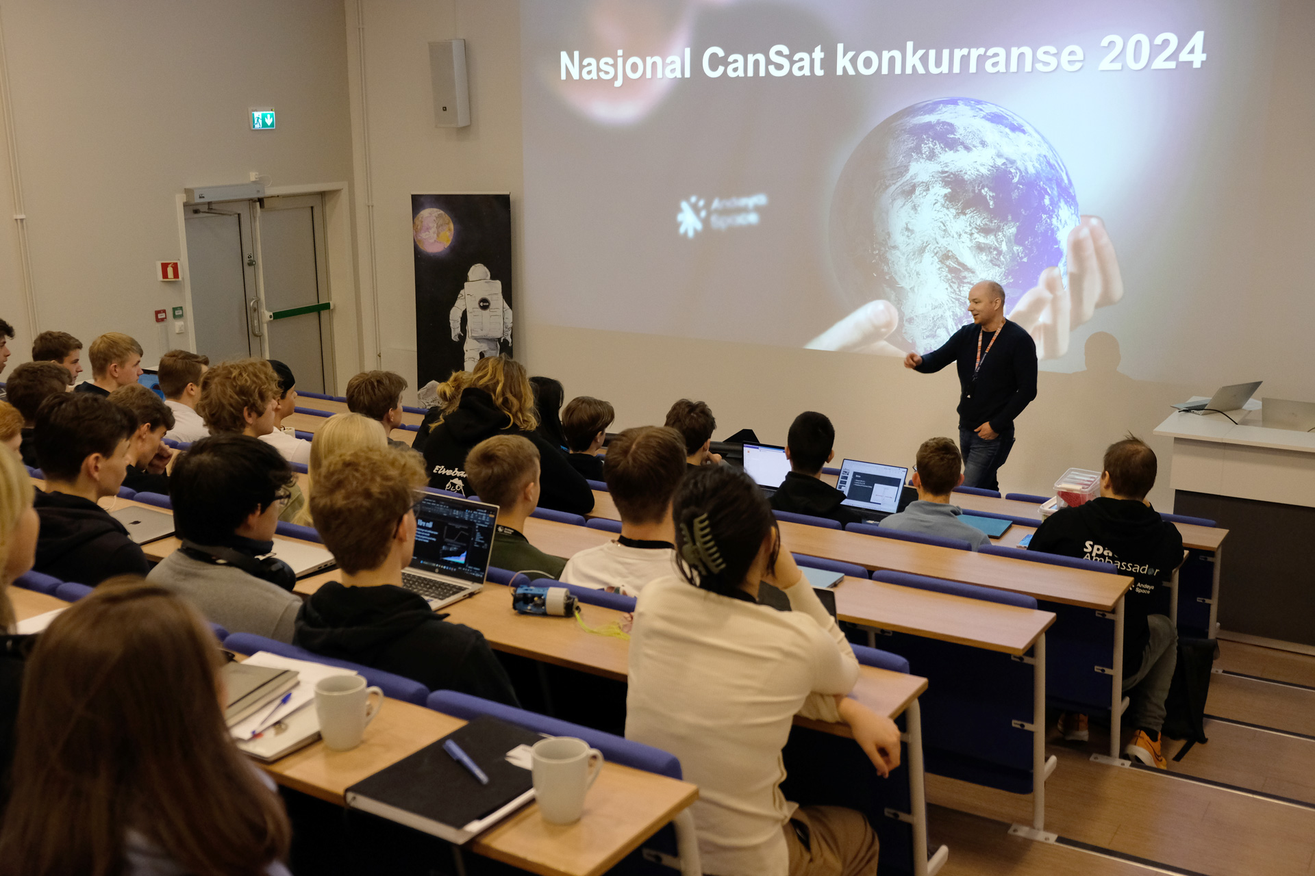 En person foredrar foran et auditorium med ungdommer. På veggen vises en presentasjon med teksten "Nasjonal CanSat konkurranse 2024".