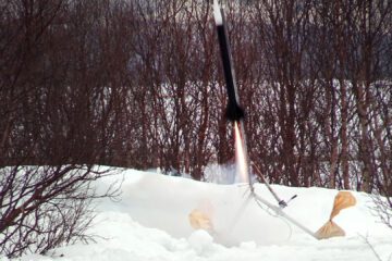En rakett blir skutt opp fra en rampe utendørs med trær og snø i for- og bakgrunn.