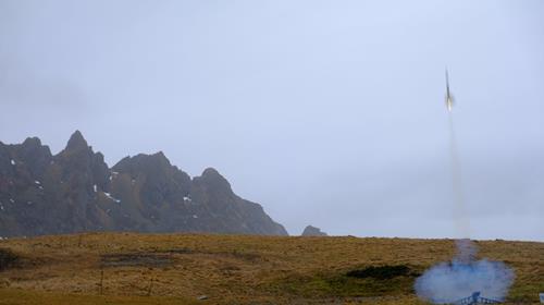 En liten modellrakett som skytes opp utendørs. Overskyet og fjell i bakgrunnen.