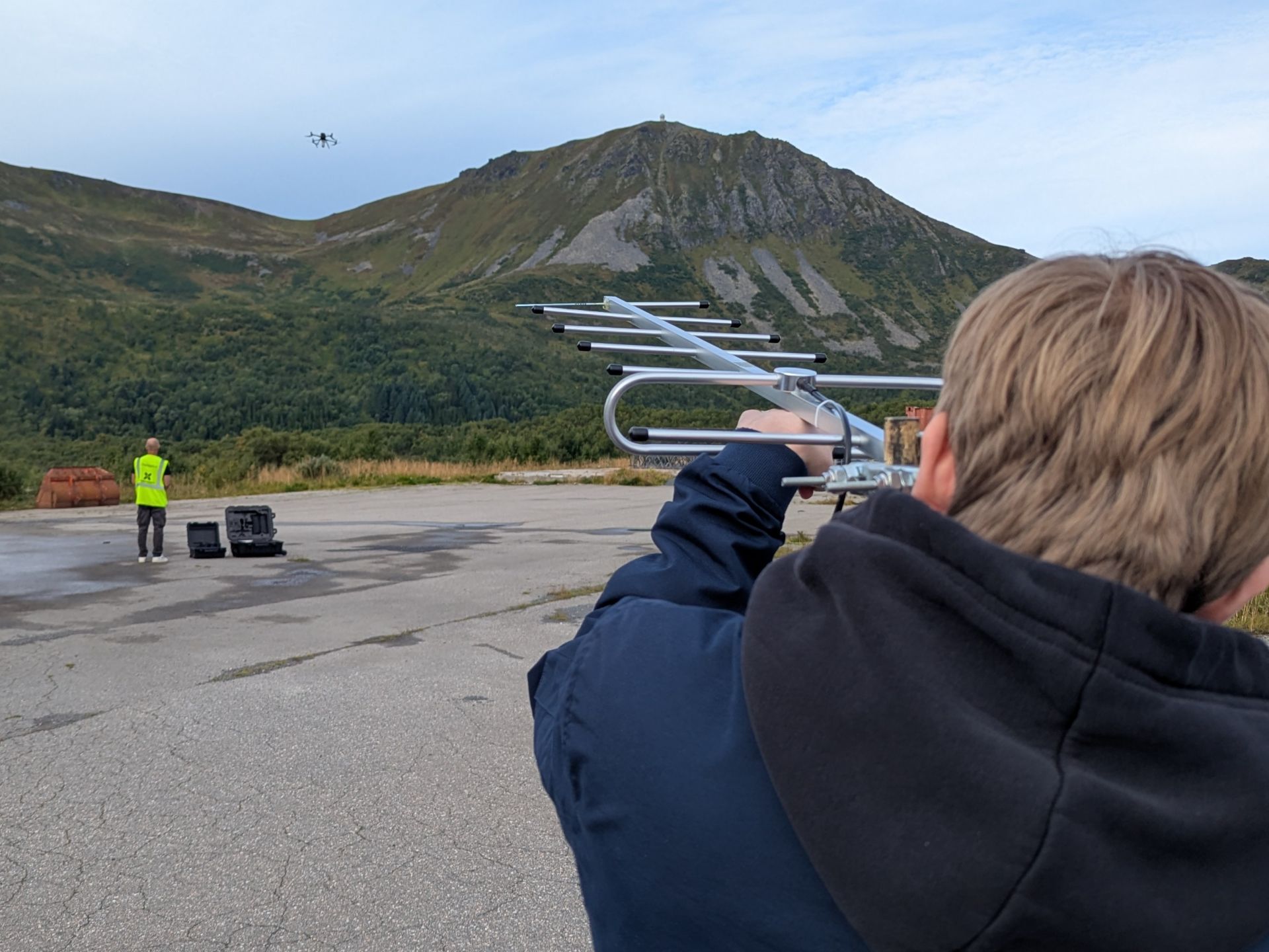 En person som holder en antenne og peker mot en drone som flyver over bakken. I bakgrunnen vises fjell og blå himmel.