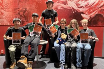 Seks smilende ungdommer som sitter og står foran en rød vegg og holder diplomer foran seg. En liten teddybjørn sitter på et av ungdommens fang.