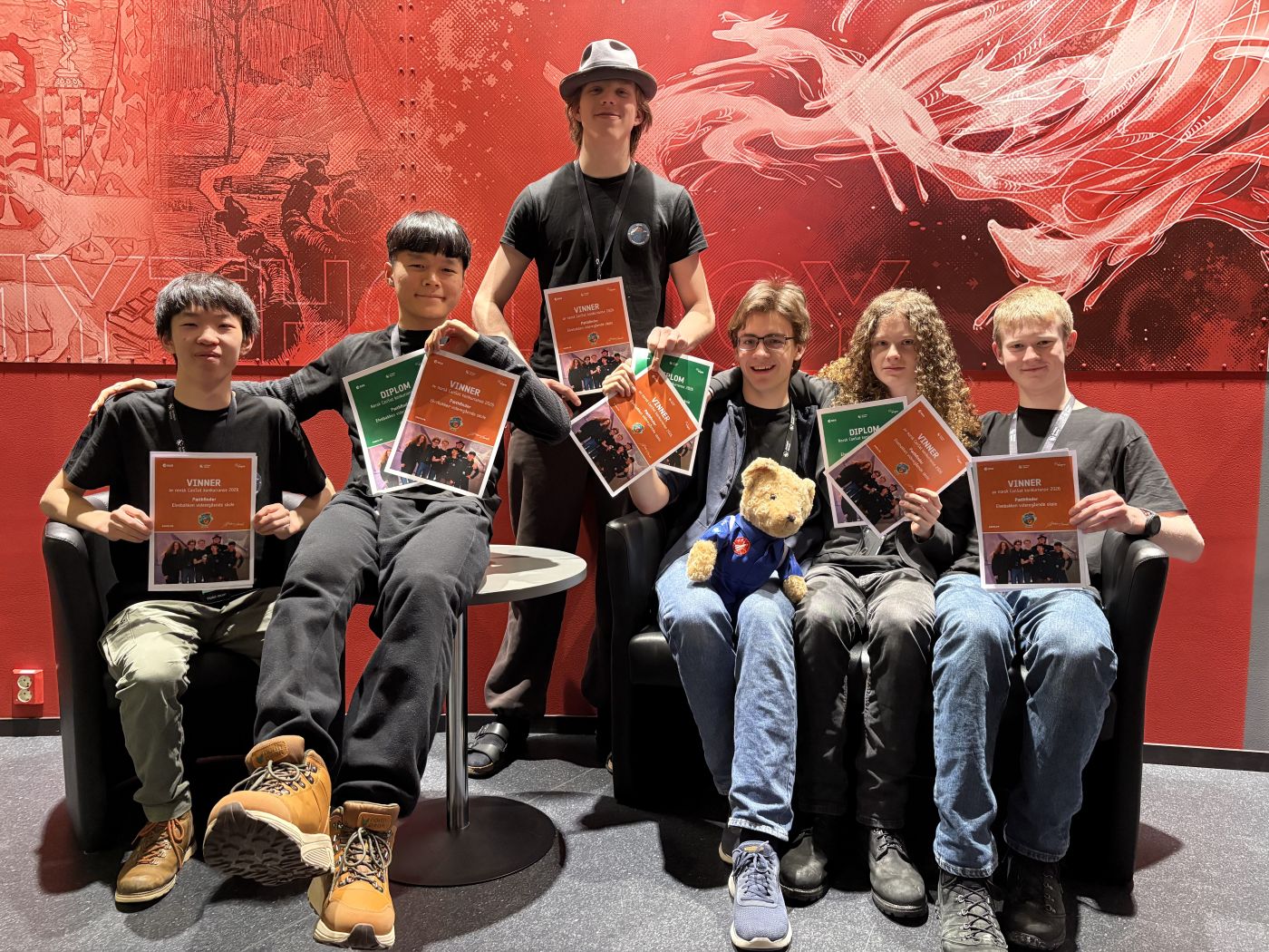 Seks smilende ungdommer som sitter og står foran en rød vegg og holder diplomer foran seg. En liten teddybjørn sitter på et av ungdommens fang.