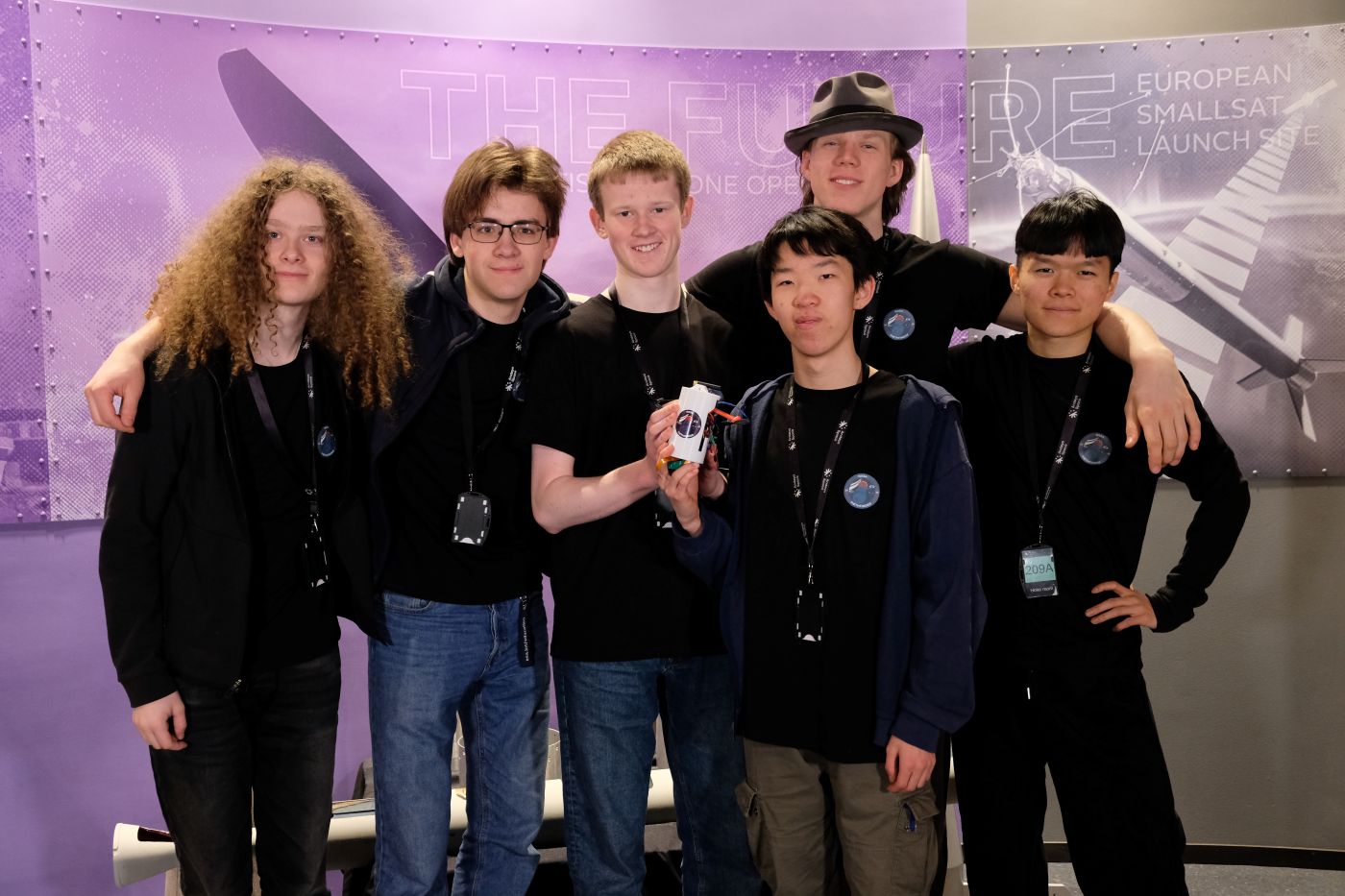 Gruppe skoleelever står samlet foran en utstillingsvegg på Andøya Space, hvor én holder en CanSat‑miniatyrsatellitt i hånden.