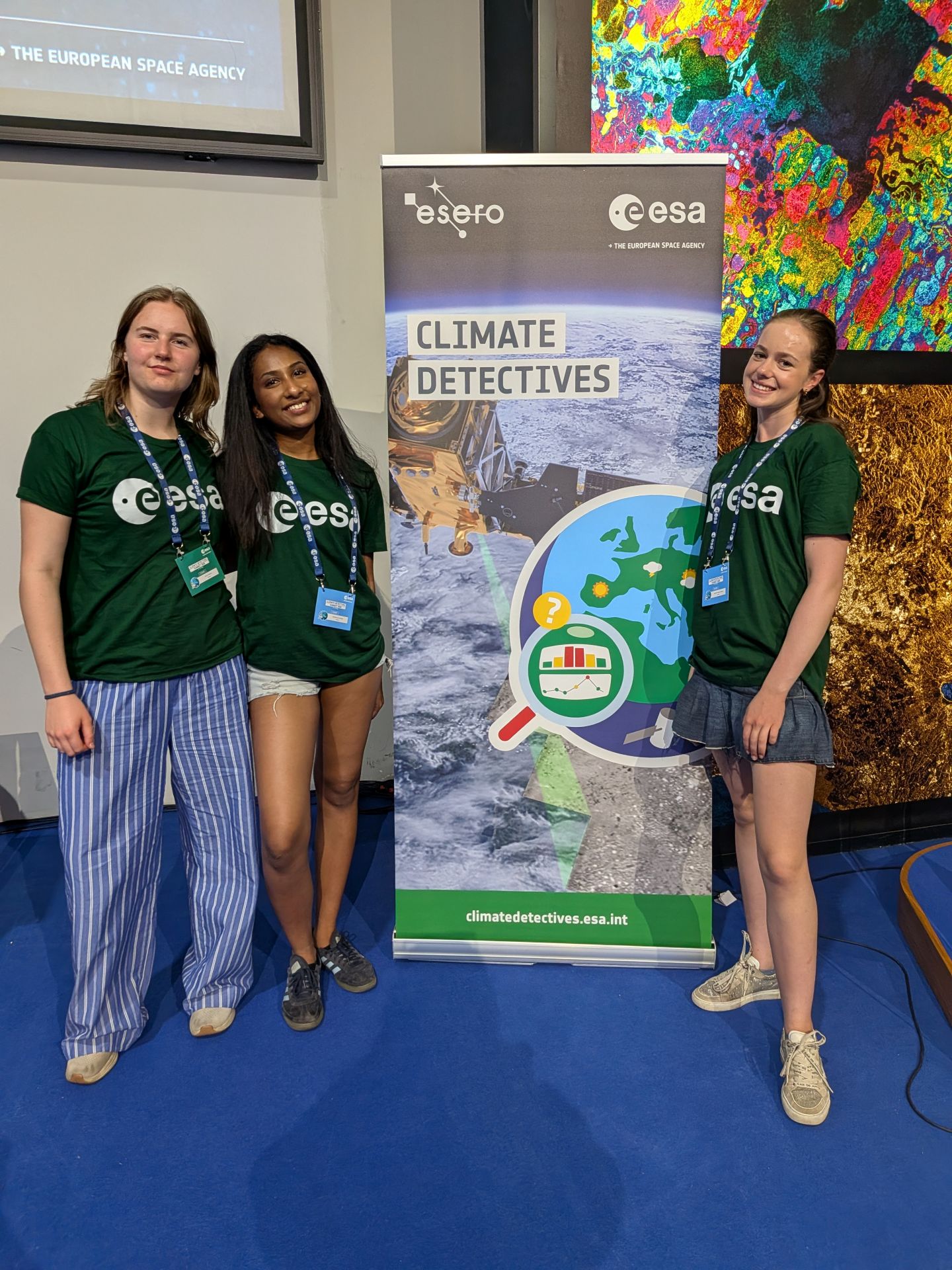 Tre ungdommer i grønne T-skjorter står ved en rollup for «Climate Detectives» fra ESA i et innendørs utstillingsområde.